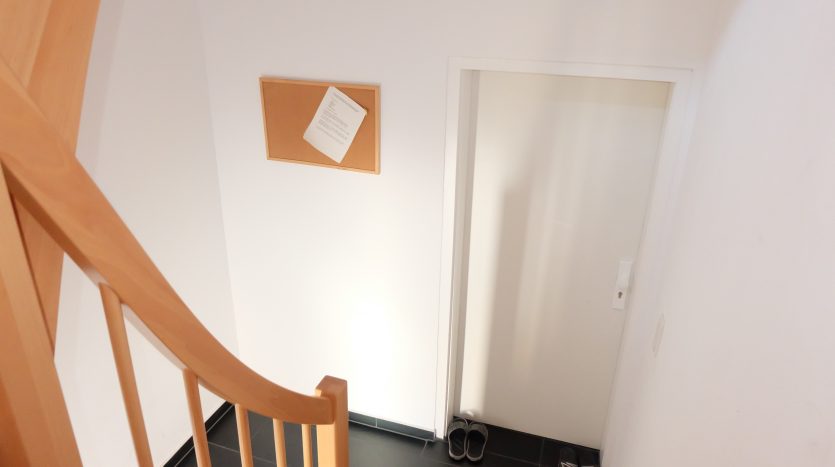 Treppenaufgang mit Holzgeländer, Tür im Hintergrund und Hausschuhen auf dem schwarzen Fliesenboden.