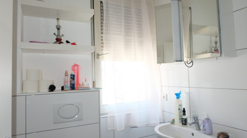 Helles Badezimmer mit Waschbecken, Spiegel, Regalen und Fenster. Diverse Pflegeprodukte auf dem Waschbecken und Regalen.