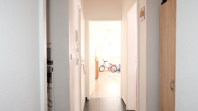 Heller Flur mit weißen Wänden, schwarzem Boden und einem rosa Kinderfahrrad im Hintergrund.