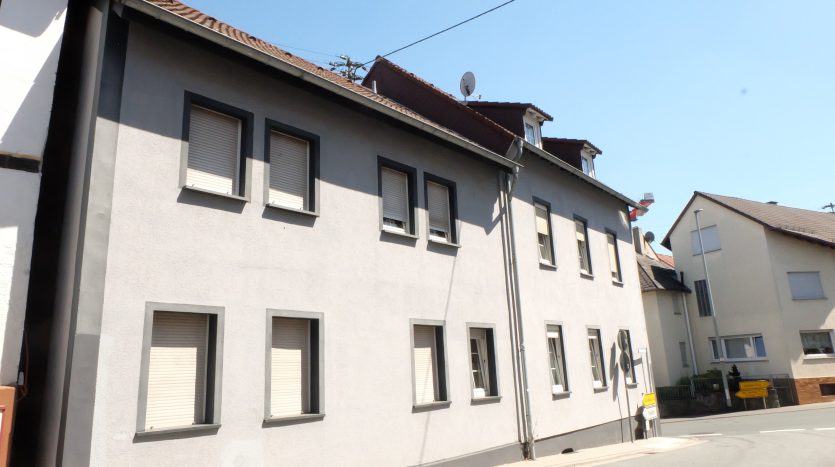 Zweistöckiges Wohngebäude mit grauer Fassade und weißen Fenstern, daneben weitere Häuser und eine Straße.