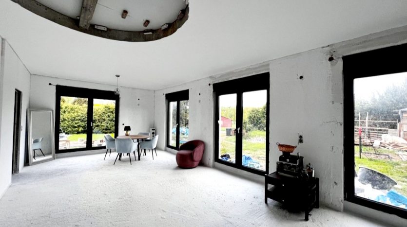 Heller, leerer Raum mit großen Fenstern, runder Deckenlampe, Tisch und Stühlen sowie einem roten Sessel.
