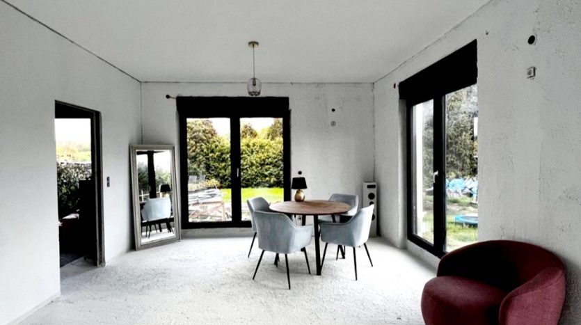 Heller Raum mit weißen Wänden, einem runden Tisch, vier blauen Stühlen und einem roten Sessel. Große Fenster bieten Ausblick.