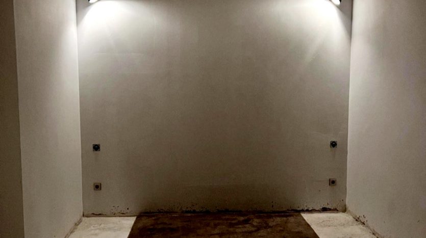Leerer Raum mit weißer Wand, zwei Wandleuchten und unebenen, dunklen Bodenflächen.