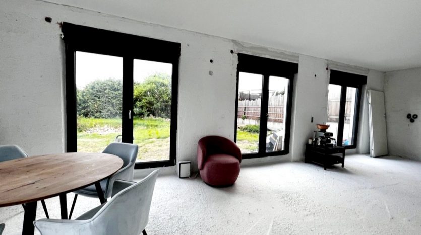 Helles, modernes Zimmer mit großen Fenstern, runder Holztisch und grauen Stühlen, rotem Sessel und weißem Boden.