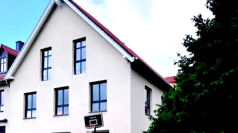Haus mit Satteldach, mehreren Fenstern und Basketballkorb im Vordergrund, umgeben von Bäumen.