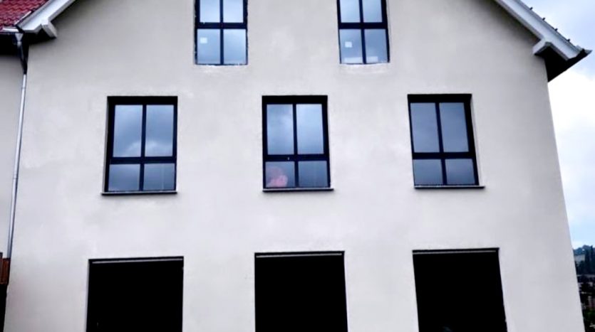 Fassade eines unvollendeten Hauses mit fünf Fenstern, unebener Erde und hohem Gras im Vordergrund.