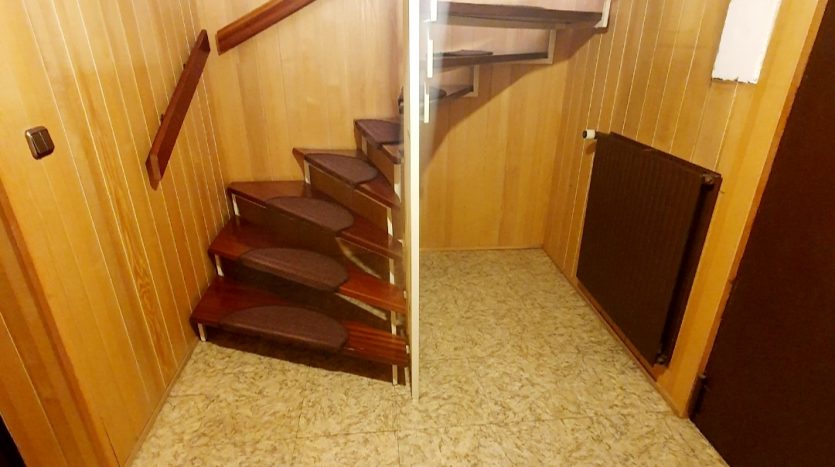 Holzverkleidete Treppe mit Holzgeländer in einem Raum mit linoleumartigem Bodenbelag und Heizkörper.