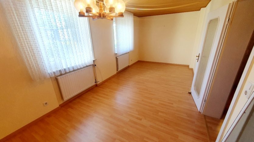 Helles Zimmer mit Holzfußboden, großen Fenstern und einer modernen Deckenlampe.