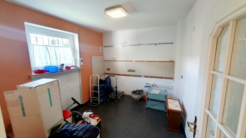 Unordentlicher Raum mit Kisten, einem Rollwagen, einem Tisch und einem Fenster mit Vorhang. Wände teilweise unstrukturiert.