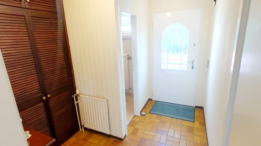 Heller Flur mit braunem Fliesenboden, Wandschrank, Heizkörper und Eingangstür mit Fenster.
