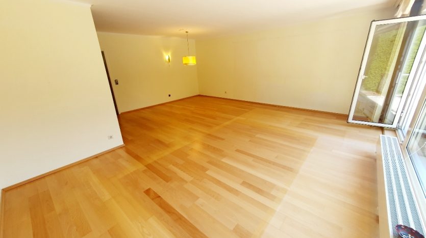 Heller, großzügiger Raum mit hellen Holzfußboden, großen Fenstern und moderner Deckenlampe.