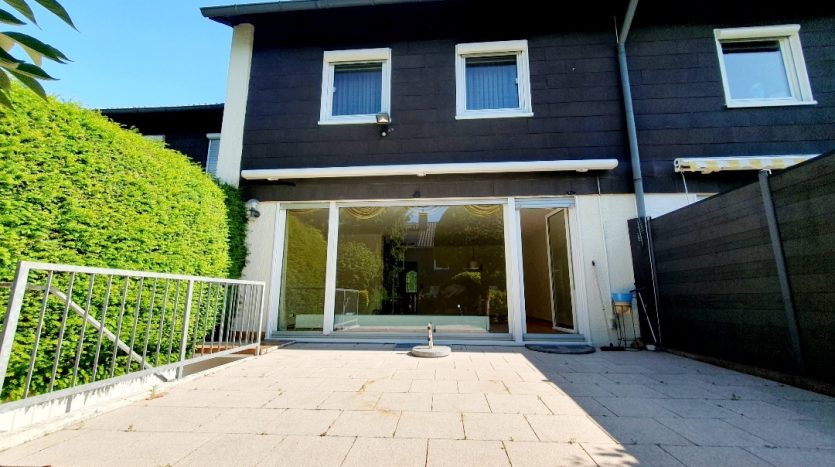 Hinteransicht eines Hauses mit großen Fenstern, Terrassenfläche und gepflegtem Grün. Klare, sonnige Atmosphäre.