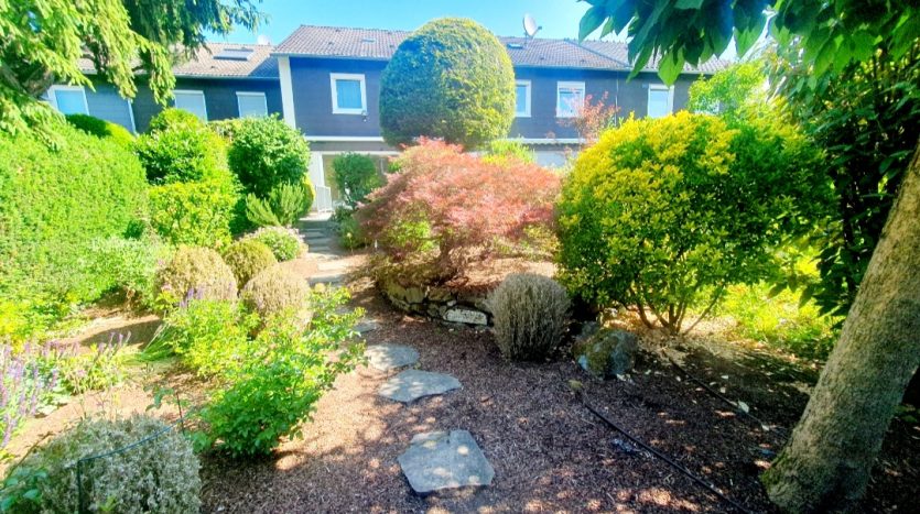 Gartenweg mit Steinplatten, umgeben von grünen Sträuchern und bunten Pflanzen, im Hintergrund ein Wohnhaus.