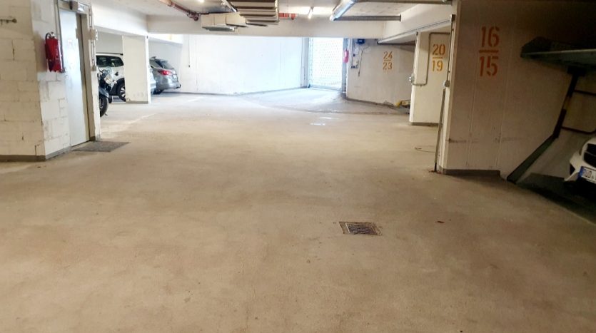 Leere Tiefgarage mit hellen Wänden, sichtbaren Parkplätzen und einer Notausgangstür.