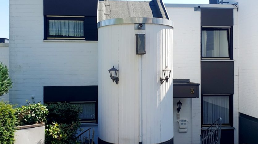 Hausfassade mit runder Erkerstruktur, weißen Wänden, schwarzen Fensterläden und gepflastertem Zufahrtsweg.