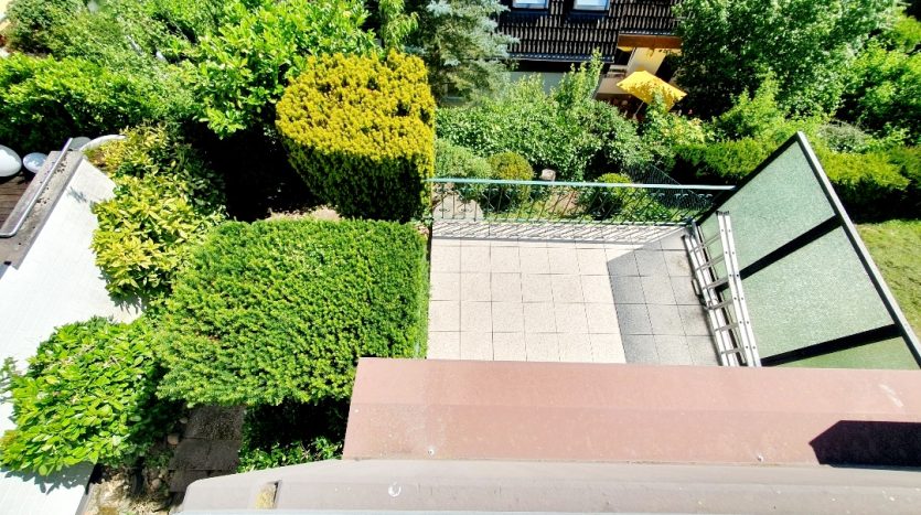 Blick von oben auf einen gepflegten Garten mit Hecken, Terrassenplatten und einem Balkon. Grüne Pflanzen und Bäume dominieren das Bild.