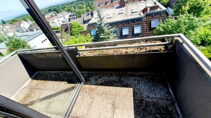 Balkon mit Sicht auf Dächer und Bäume, offenes Fenster, sonniger Tag, graue Bodenfliesen, bepflanzter Balkonkasten.
