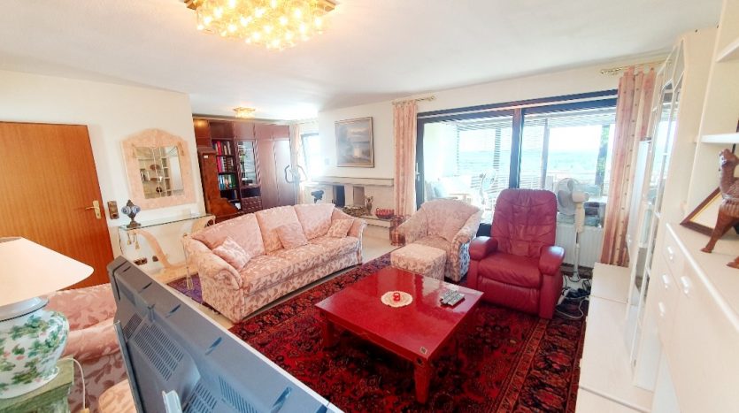 Heller Wohnraum mit zwei beigen Sofas, einem roten Tisch, einem roten Sessel und großen Fenstern.