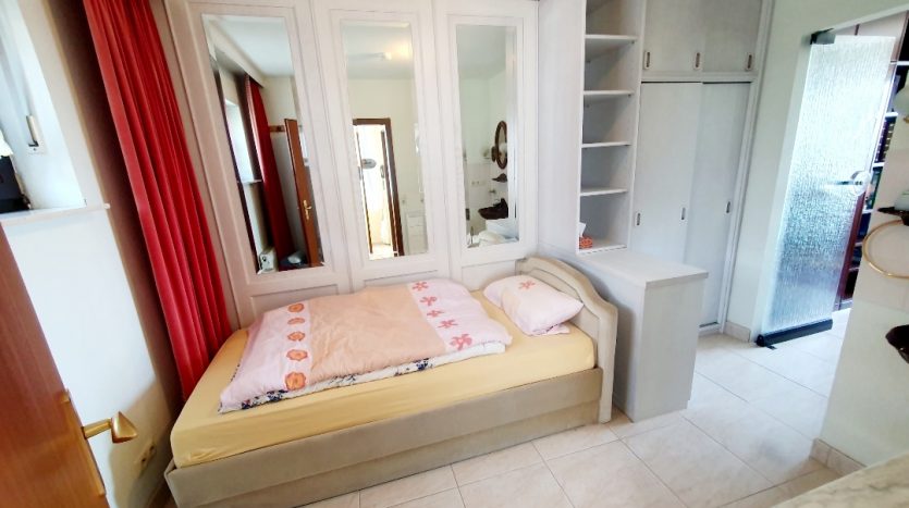 Kleines Schlafzimmer mit einem Bett, Spiegelkleiderschrank, Regal und roten Vorhängen, helle Fliesen.
