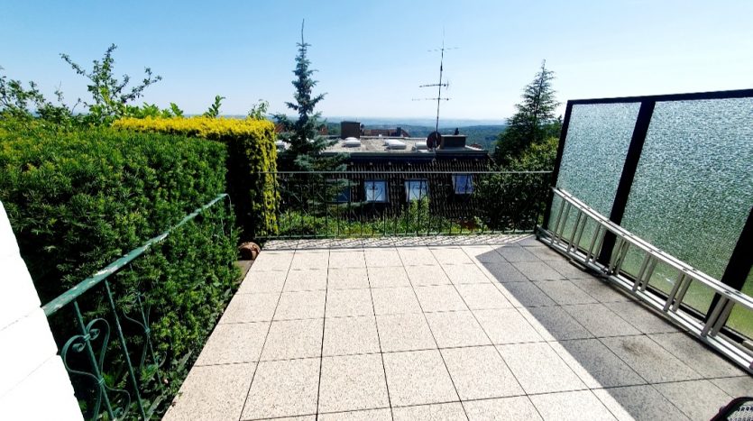 Terrasse mit Fliesen, umgeben von grünen Hecken und Blick auf die Landschaft, Sonnenschein, metallene Geländer.