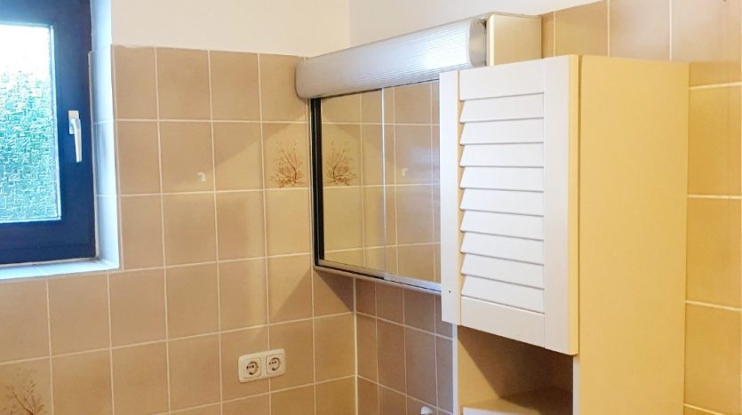 Kleines Badezimmer mit einem runden, rosa Waschbecken, Spiegel und einem weißen Schrank. Fliesen in Beige.