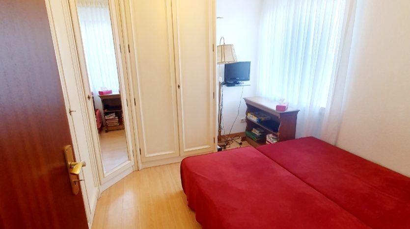 Kleines Schlafzimmer mit roten Betten, einem Kleiderschrank, TV und großen Fenstern mit Vorhängen.