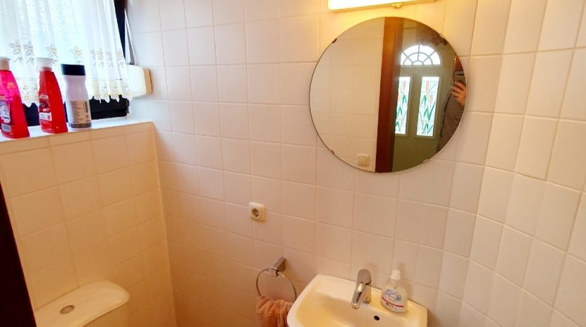 Kleines Badezimmer mit WC, Waschbecken, Spiegel und Abfalleimer. Helle Fliesen und Fenster mit Vorhängen.