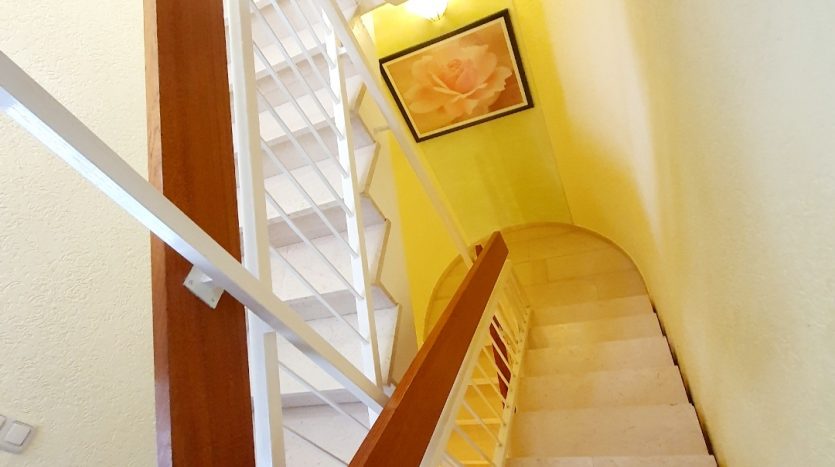 Treppenhaus mit gelben Wänden, weißen Treppenstufen und Holzgeländer, Blick nach unten auf die Treppe.
