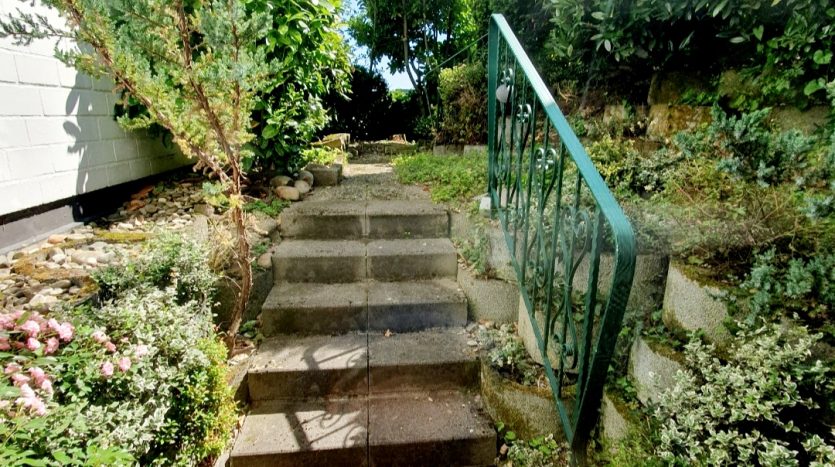 Zwei Reihen Steinstufen führen durch einen bepflanzten Garten mit Blumen und Büschen zu einem grünen Geländer.