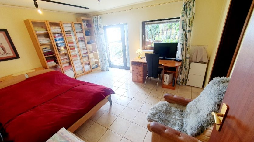 Helles Schlafzimmer mit rotem Bett, Schreibtisch, Computer, Sessel und Regal mit Handtüchern. Zugang zur Terrasse.