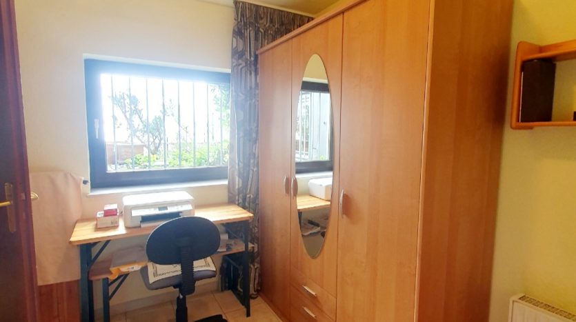 Helles Zimmer mit Holzschrank, Schreibtisch und Stuhl. Fenster mit Blick ins Freie, Fliesenboden.