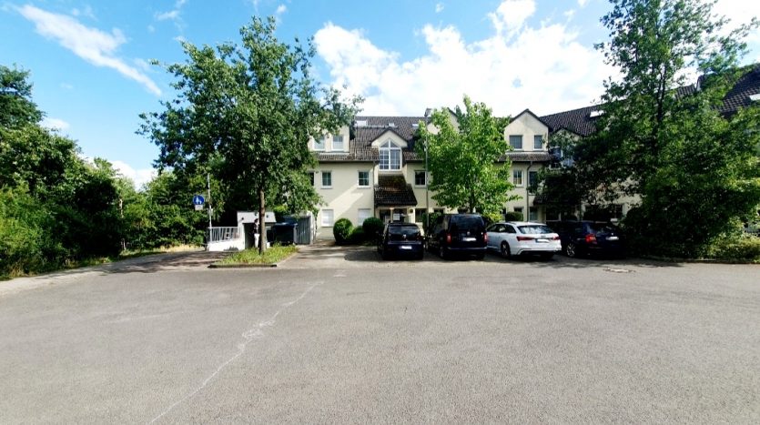 Parkplatz vor einem mehrstöckigen Wohngebäude, umgeben von Bäumen und blauem Himmel, mehrere Autos geparkt.