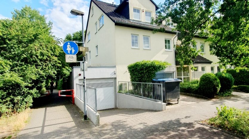 Zwei- bis dreigeschossiges Wohnhaus mit hellem Putz, blauen Mülltonnen und Fußgängerweg mit Schild.