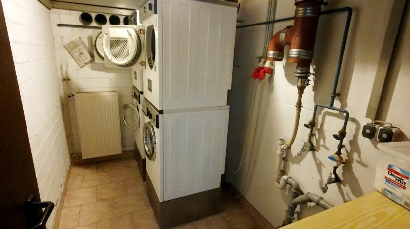Waschküche mit Waschmaschine und Trockner, Heizkörper, Wasserrohren und Fliesenboden. Deckenbeleuchtung vorhanden.