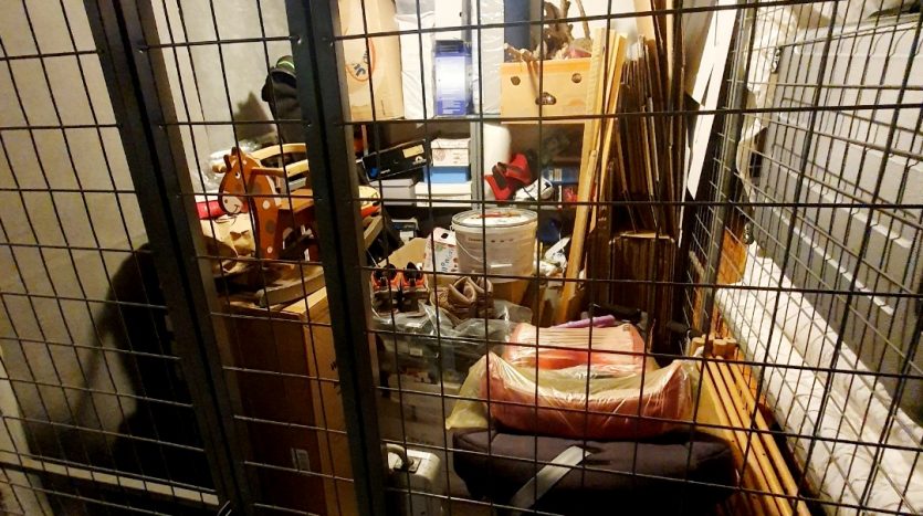 Abgetrennter Lagerraum mit Gittertür, voll mit Kartons, Möbeln und verschiedenen Gegenständen. Beleuchtet von einer Deckenlampe.