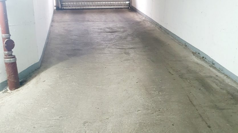 Blick auf einen langen, leeren, betonierten Flur mit einer geschlossenen Rolltür am Ende und Rohren an der Wand.