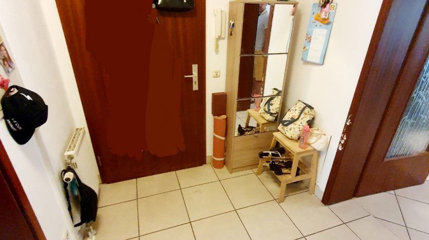 Flur mit brauner Tür, Spiegel, Schuhregal und Taschen. Fliesenboden und Heizkörper sichtbar.