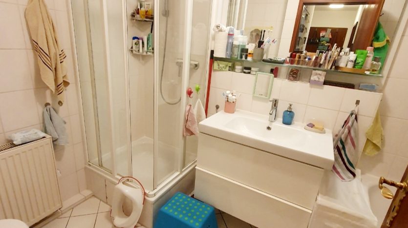 Kleines Badezimmer mit Dusche, Waschbecken, Toilette und blauer Fußbank. Weiße Fliesen und Spiegelschrank sichtbar.