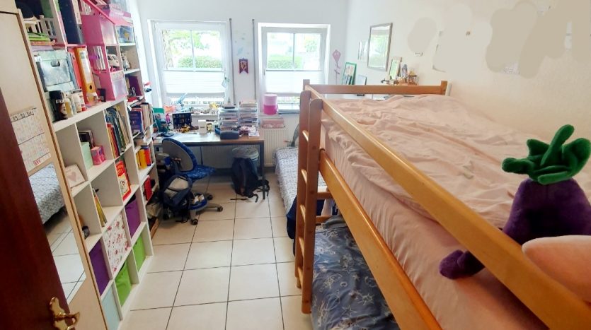 Kleines Zimmer mit Etagenbett, Bücherregal, Schreibtisch und zwei Fenstern. Helle Fliesen und bunte Dekoration.