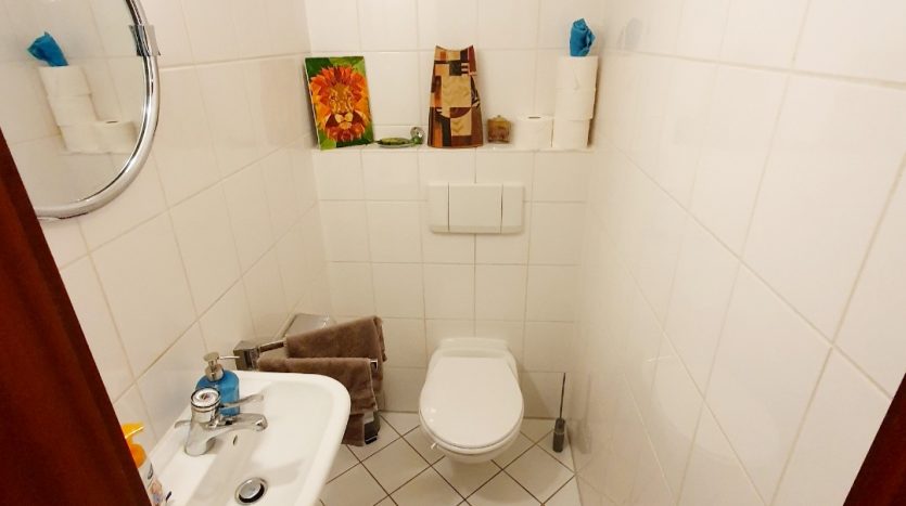 Kleines, helles Badezimmer mit weißen Fliesen, Waschbecken, WC und Holzregal. Dekorative Bilder an der Wand.