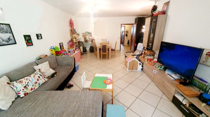 Wohnzimmer mit grauem Sofa, Tisch, Spielzeug und TV. Helle Wände, Fliesenboden, gemütliche Einrichtung.