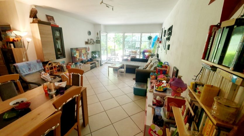 Heller Wohnraum mit Essbereich, Couch, Fernseher und Spielzeug. Große Fenster bieten Zugang zu einer Terrasse.