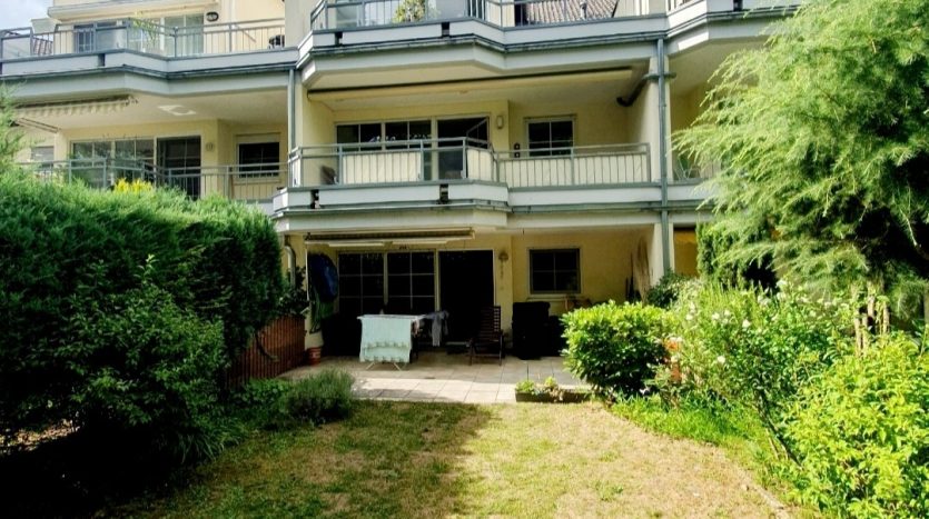 Drei moderne Mehrfamilienhäuser mit Balkonen, umgeben von einem gepflegten Garten und blühenden Pflanzen.