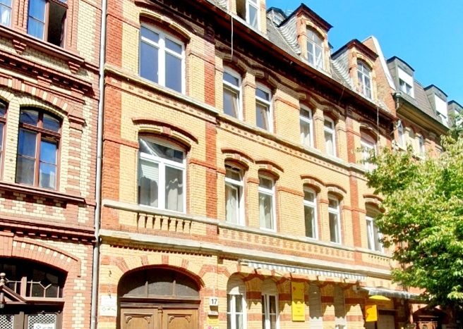 Historisches Backsteingebäude mit mehreren Fenstern, großer Holztür und einem grauen Auto auf der Straße.