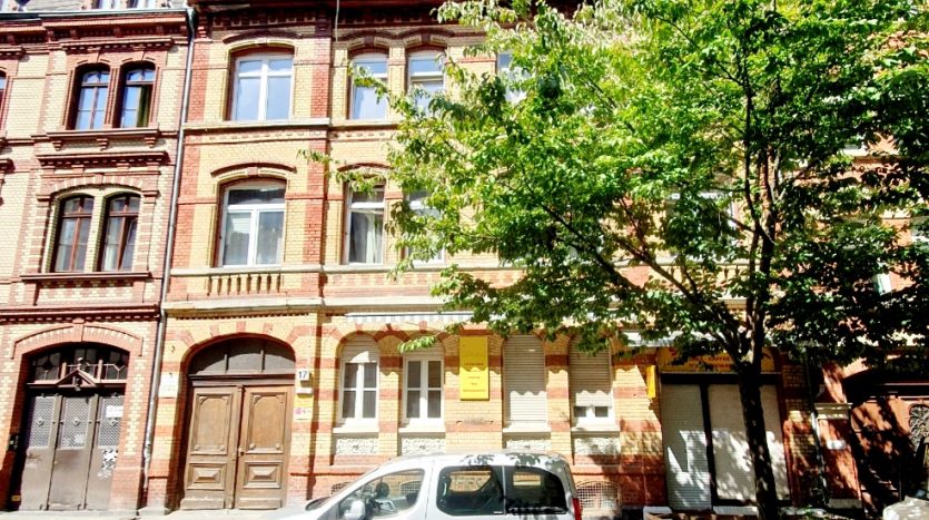 Rote Backsteinfassade mit großen Fenstern, im Vordergrund ein grauer Kleinwagen, umgeben von Bäumen.