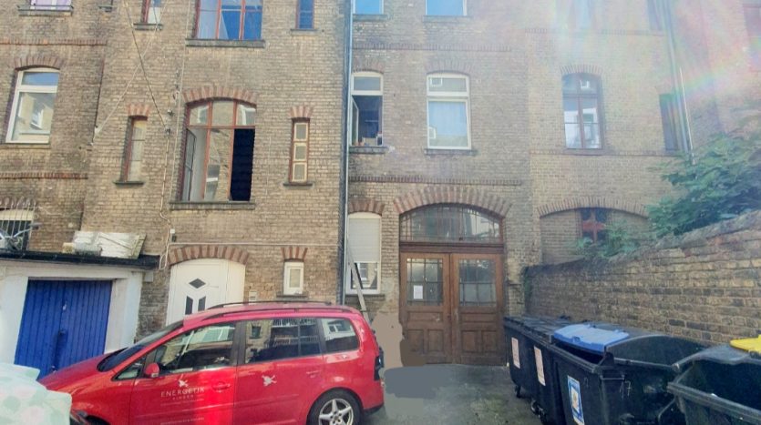 Hinterhof mit Backsteinfassade, rotem Auto und Mülltonnen. Wäsche hängt vor einem Fenster. Tageslicht und Schatten.