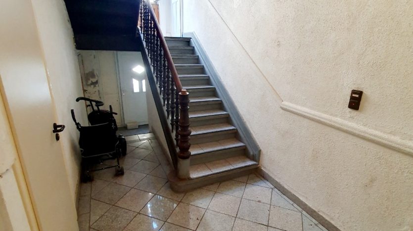 Heller Treppenhausbereich mit Holztreppe, Fliesenboden und einem Rollstuhl in der Ecke.
