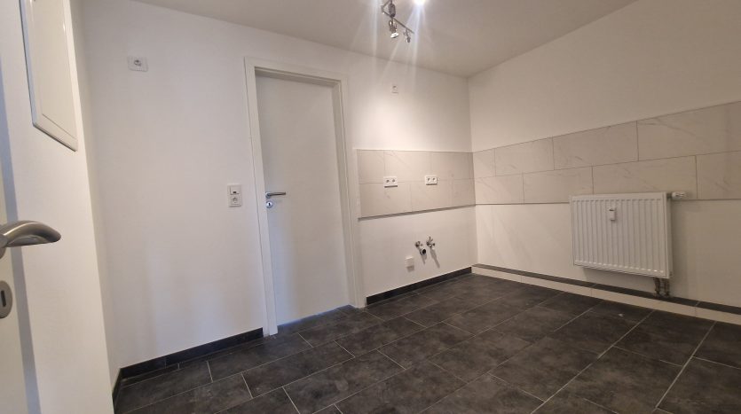 Heller Raum mit dunklem Fliesenboden, weißer Wand, Heizkörper und Tür. Deckenstrahler sorgen für Beleuchtung.