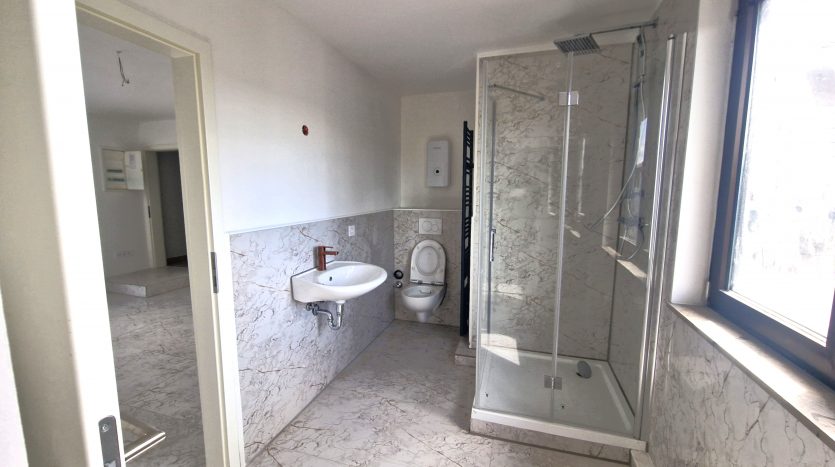 Helles Badezimmer mit Dusche, WC, Waschbecken und marmoriertem Bodenbelag. Fenster sorgt für Tageslicht.