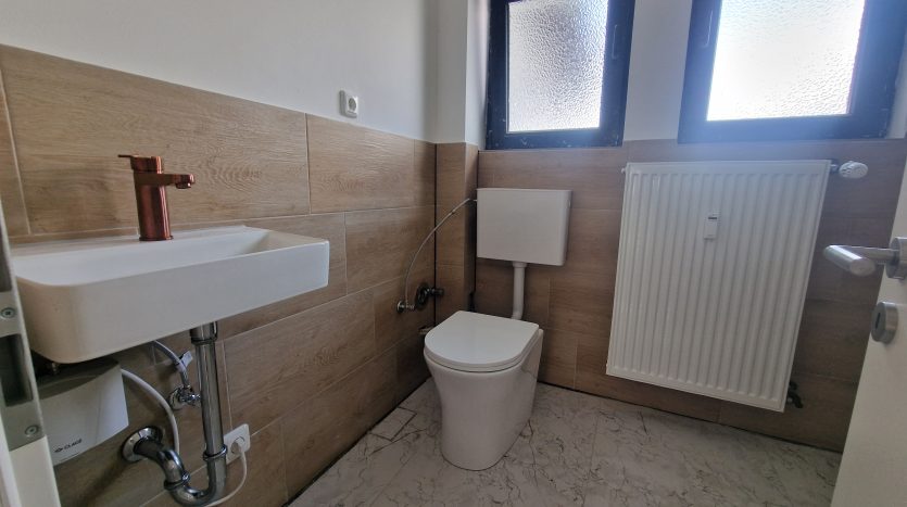 Kleines Badezimmer mit weißen Fliesen, Wandwaschbecken und WC, Kupferarmatur und zwei Fenstern.